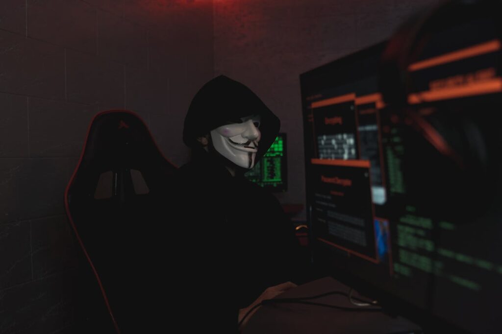 An anonymous hacker wearing a mask working on a computer in a dark room.